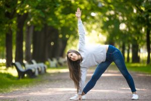 Smiling beautiful sporty young woman working out in park alley, doing stretching exercise, standing in Triangle yoga pose, Trikonasana, full length