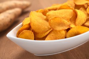 Crispy Peruvian sweet potato chips in white ceramic bowl with sweet potatoes in the back (Selective Focus, Focus one third into the sweet potato chips)