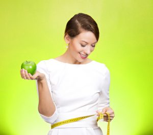 Closeup portrait young attractive, happy, fit woman in white dress, holding lime apple, measuring her waist, isolated green background. Healthy life style, nutrition. Positive emotion, expression