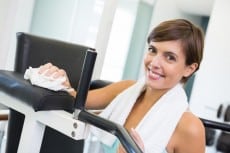 Fit brunette wiping down bench smiling at camera at the gym